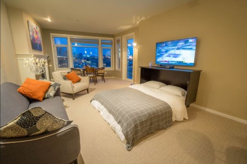 Cozy studio apartment with a bed, sofa, TV, and dining area, featuring large windows with a city view.