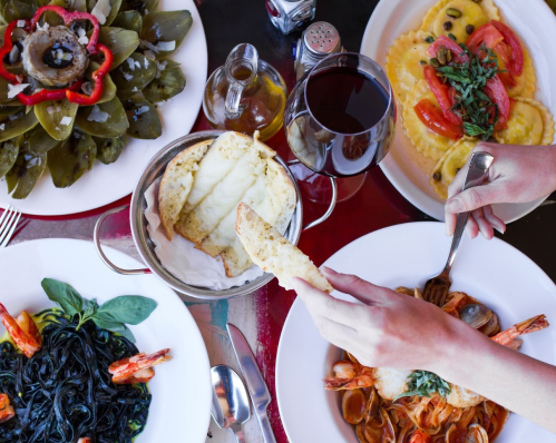 A table set with various dishes, including pasta, seafood, and a salad, with a hand reaching for bread.