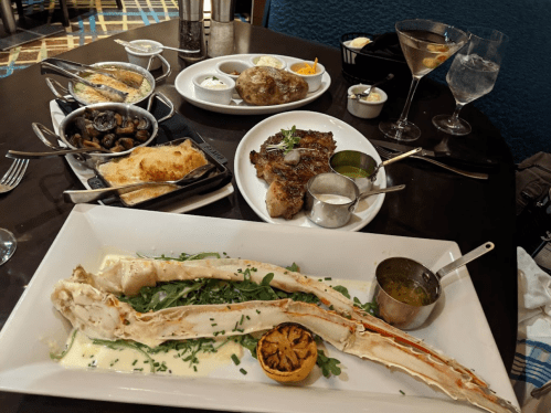A table set with a variety of dishes, including crab legs, steak, sides, and drinks, in a restaurant setting.