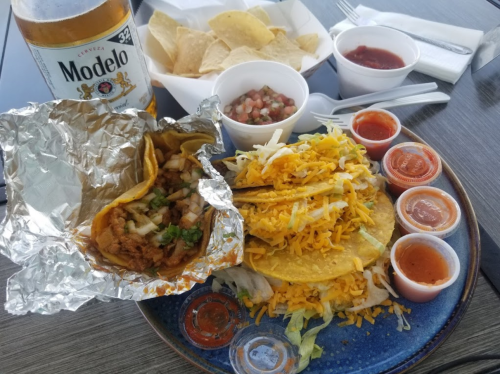 A plate of tacos with cheese, lettuce, and sauces, accompanied by tortilla chips, salsa, and a beer.