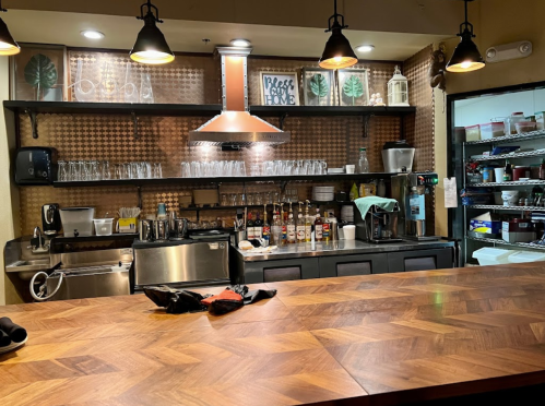 A cozy café counter with a wooden surface, shelves of glassware, and various coffee supplies in the background.