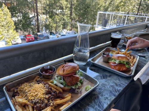 Two burgers with fries on a table, accompanied by a glass of water and a dark beverage, set against a forest backdrop.