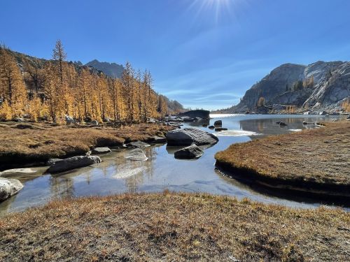 A serene landscape featuring a calm lake, rocky shore, and golden autumn trees under a clear blue sky.