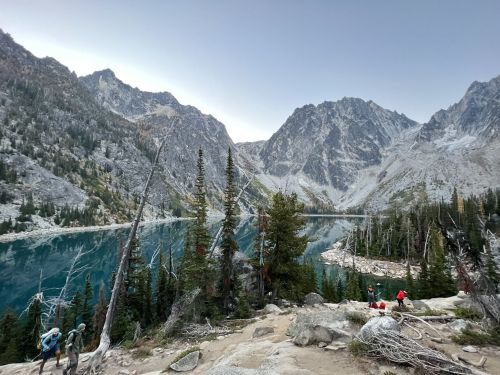 A scenic view of a mountain lake surrounded by tall trees and rugged mountains, with hikers in the foreground.