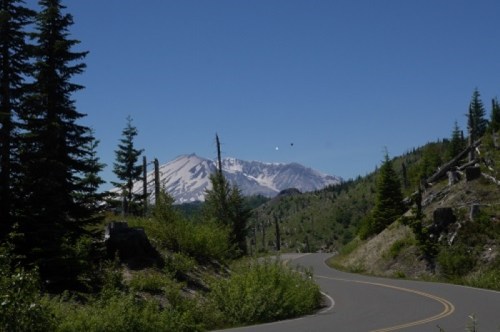 A winding road through green trees with a view of snow-capped mountains under a clear blue sky.