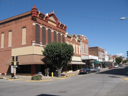 Historic buildings line a street in a small town, featuring brick facades and awnings under a clear blue sky.