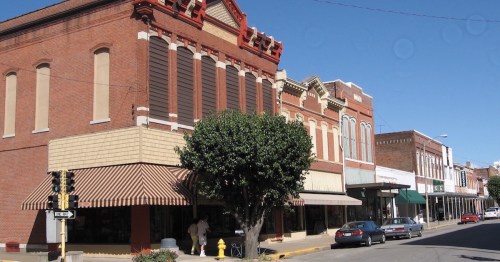 Historic brick buildings line a sunny street, featuring awnings and greenery, with parked cars and clear blue skies.