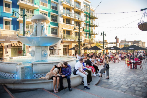 A lively plaza with a fountain, couples, and groups enjoying outdoor seating at cafes and restaurants.
