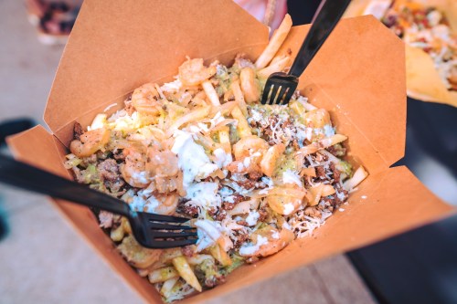 A takeout box filled with shrimp, beef, fries, and drizzled with sauce, served with two black forks.