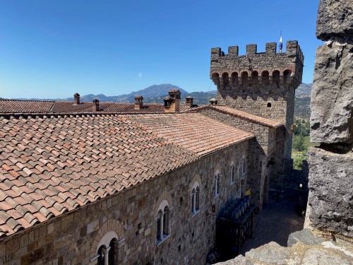 A stone castle with a tiled roof and tower, set against a clear blue sky and distant mountains.