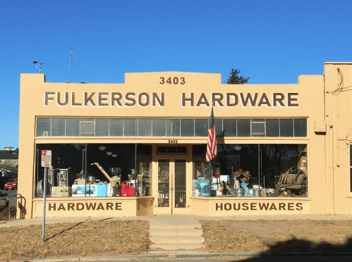 A peach-colored hardware store with large windows displaying tools and housewares, featuring an American flag out front.