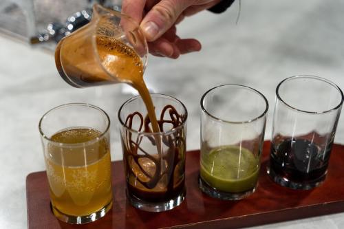 A hand pours a dark liquid into a glass, surrounded by three other glasses with different colored liquids on a wooden tray.