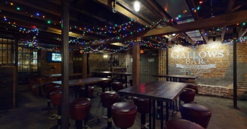 A dimly lit underground bar with high-top tables, leather stools, string lights, and "The Gallows Bar" painted on brick.