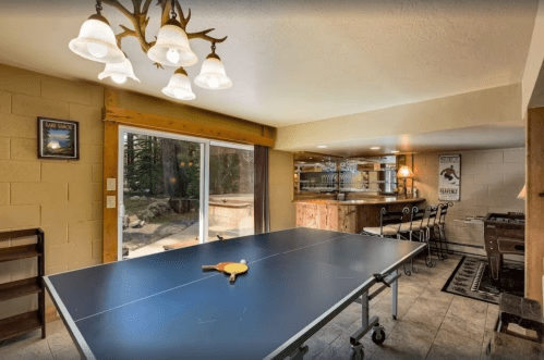 A spacious game room featuring a ping pong table, bar area, and large windows with a view of the outdoors.