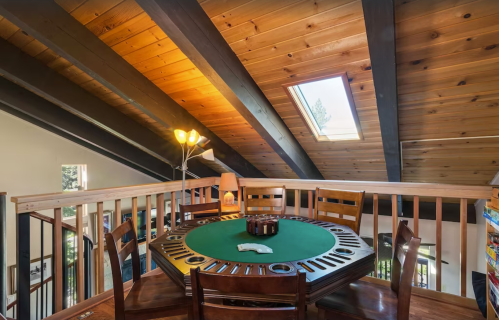 A cozy game room with a green felt table, wooden beams, and a skylight, surrounded by chairs and warm lighting.