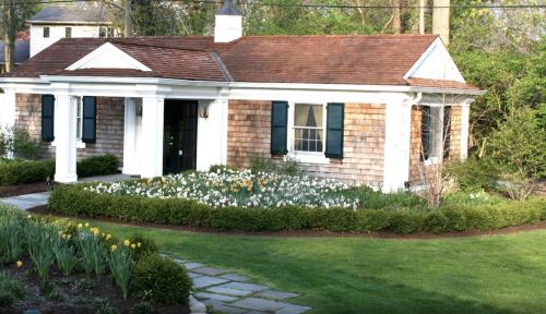 A charming house with a shingled exterior, white trim, and a flower garden in front, surrounded by green grass.