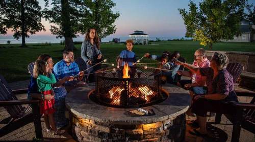 A group of people roasting marshmallows around a fire pit at sunset, with a lake and gazebo in the background.