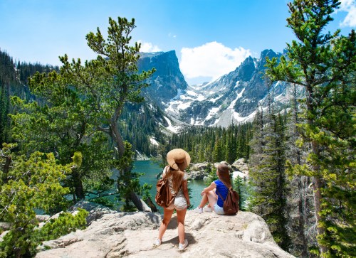 Two women sit on a rock, overlooking a scenic mountain lake surrounded by trees and snow-capped peaks under a blue sky.