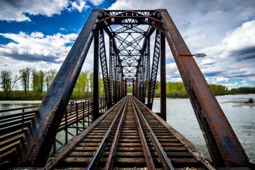 Rusty metal bridge over a river, with train tracks leading into the distance under a cloudy blue sky.