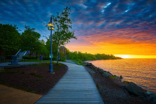 A scenic waterfront path lined with trees, leading to a vibrant sunset over the water, with clouds in the sky.
