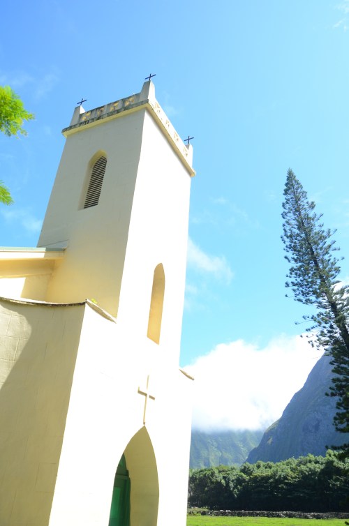 A white church tower with a cross, surrounded by lush greenery and mountains under a clear blue sky.