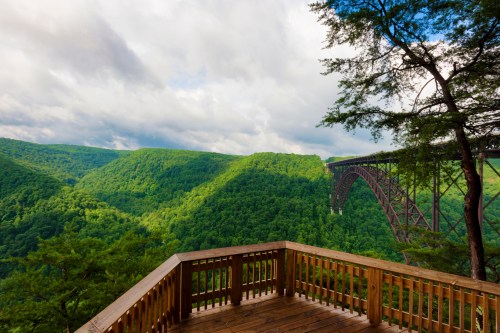 A wooden overlook with a view of lush green mountains and a large bridge spanning a deep valley under a cloudy sky.
