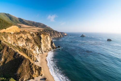 A scenic coastal view featuring cliffs, a sandy beach, and calm blue waters under a clear sky.