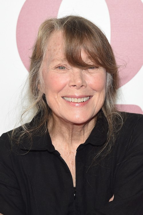 A smiling woman with long, wavy hair wearing a black shirt, posing against a light pink background.