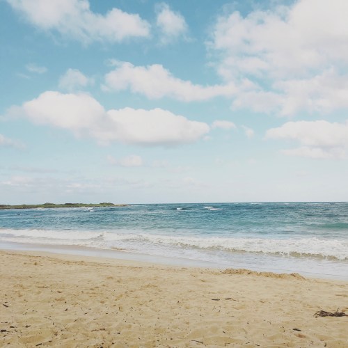 A serene beach scene with soft waves, golden sand, and a clear blue sky dotted with fluffy clouds.