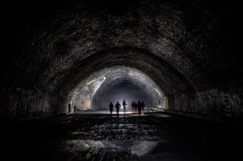 Silhouettes of people stand in a dark, arched tunnel with faint light in the background, reflecting on the wet ground.