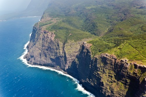 Aerial view of a rugged coastline with steep cliffs meeting a vibrant blue ocean. Lush greenery covers the land.