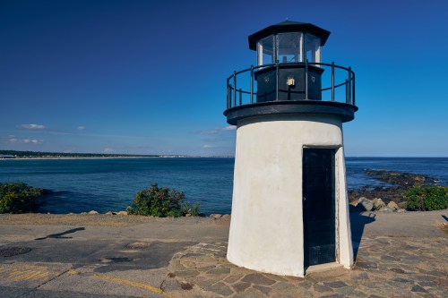 A white lighthouse with a black top stands by the shore, overlooking a calm blue sea under a clear sky.