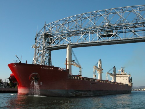 A large cargo ship passing under a steel bridge on a clear day, with blue skies and calm waters.