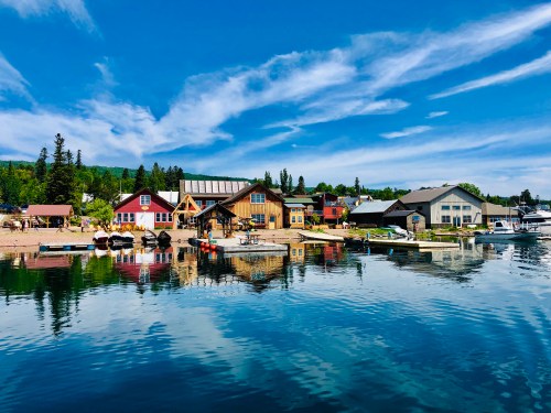 Colorful houses line a calm waterfront, reflecting in the water under a bright blue sky with scattered clouds.