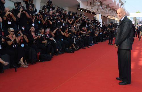 A man in a black suit stands on a red carpet, surrounded by photographers capturing the moment.