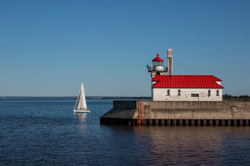 A sailboat glides past a lighthouse with a red roof on a calm blue lake under a clear sky.