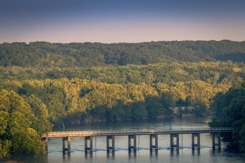 A serene river scene with a bridge crossing over calm waters, surrounded by lush green trees and hills in the background.