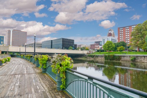 Scenic view of a riverwalk with flowers, a bridge, and city buildings under a blue sky with clouds.