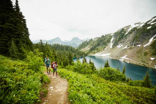 Two hikers walk along a trail surrounded by lush greenery and mountains, with a serene lake in the background.