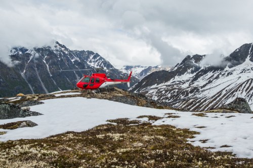 A red helicopter parked on a snowy mountain landscape with rugged peaks and cloudy skies in the background.
