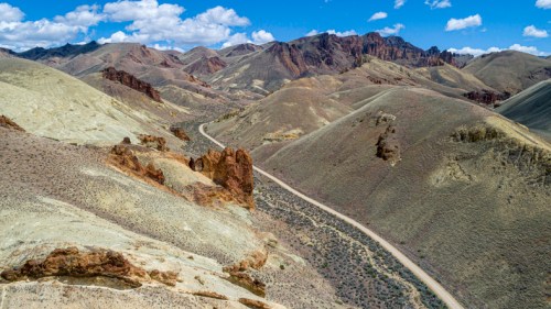 Aerial view of rugged mountains and rolling hills under a blue sky with scattered clouds, featuring a winding dirt road.
