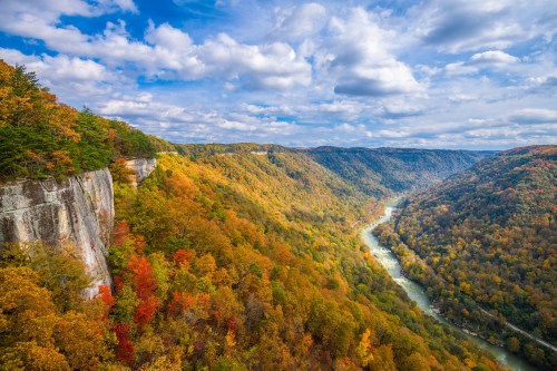 A scenic view of a river winding through colorful autumn foliage and steep cliffs under a partly cloudy sky.