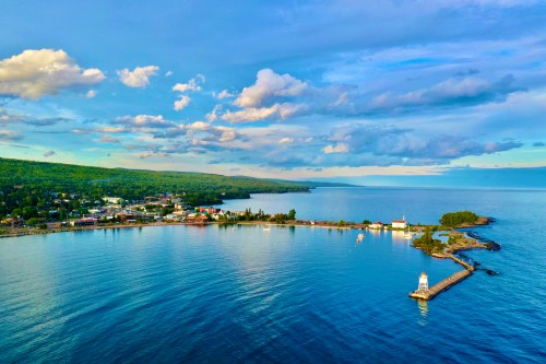 Aerial view of a coastal town with a lighthouse, calm waters, and lush green hills under a blue sky with clouds.