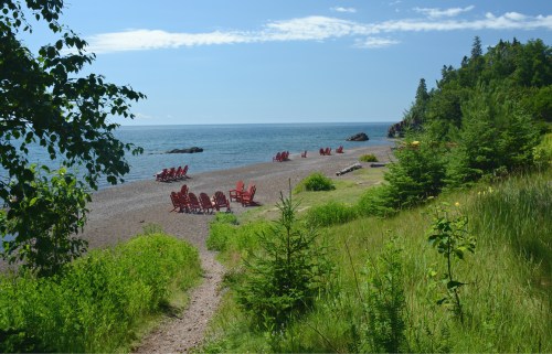 A serene beach scene with red chairs on a pebbly shore, surrounded by greenery and a calm blue ocean under a clear sky.