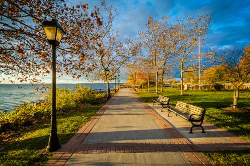 A scenic waterfront path lined with benches and trees, illuminated by a lamp under a partly cloudy sky.