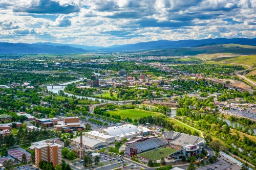 Aerial view of a vibrant city surrounded by green hills, rivers, and mountains under a partly cloudy sky.