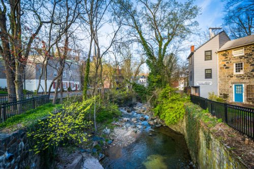 A serene stream flows through a lush green area, surrounded by trees and historic buildings under a clear blue sky.