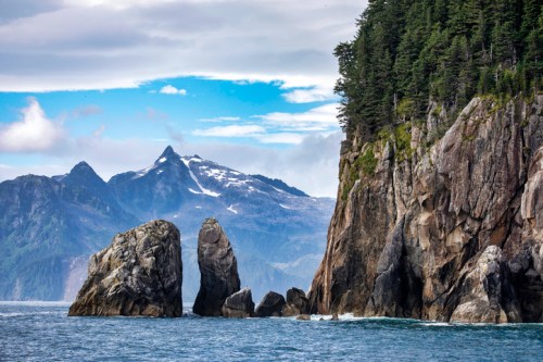 Rocky coastline with towering cliffs, lush greenery, and snow-capped mountains under a cloudy sky.