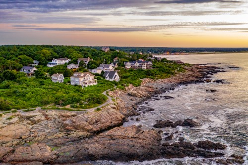 Aerial view of coastal homes on rocky cliffs at sunset, surrounded by lush greenery and calm waters.