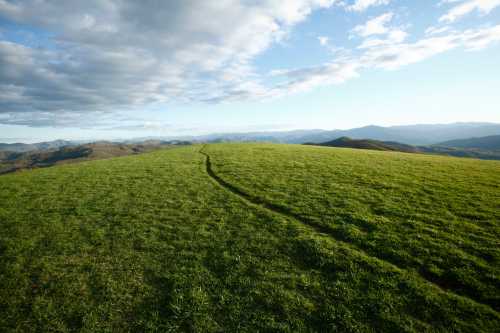 A grassy hilltop with a winding path, surrounded by distant mountains and a partly cloudy sky.